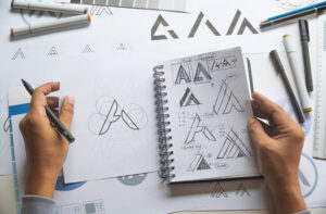 Two hands hold a notebook full of logo sketches and a pen on a desk. Ideating on potential branding and logo concept development.