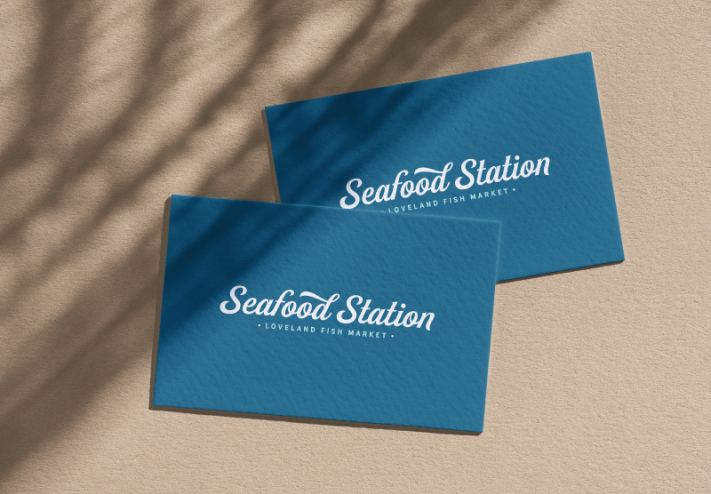An image of the Seafood Station business card design. Two navy blue cards stacked on each other on a tan background, with a shadow falling across a corner.